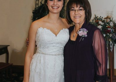 Bride and mother smiling while mother wears pin-on corsage that includes purple roses and silver ribbon.