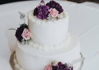 Two tired white cake that has groupings of flowers placed on the top, left side between tiers, and bottom. The flower cluster include purple roses, purple carnations, and silver eucalyptus.