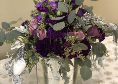 Centerpiece in a mercury silver vase with purple roses, purple stock, and eucalyptus.