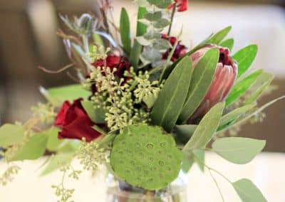 Centerpiece arrangement including lotus pods, eucalyptus, burgundy roses, protea, and burgundy roses.