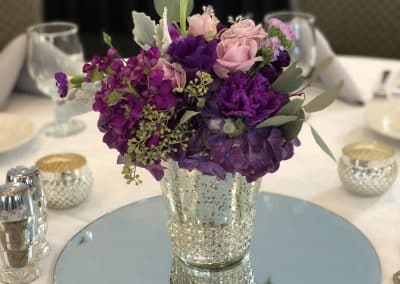 Centerpiece in a mercury silver vase with purple roses, purple stock, and eucalyptus.