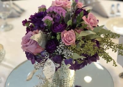 Centerpiece in a mercury silver vase with purple roses, purple stock, and eucalyptus.